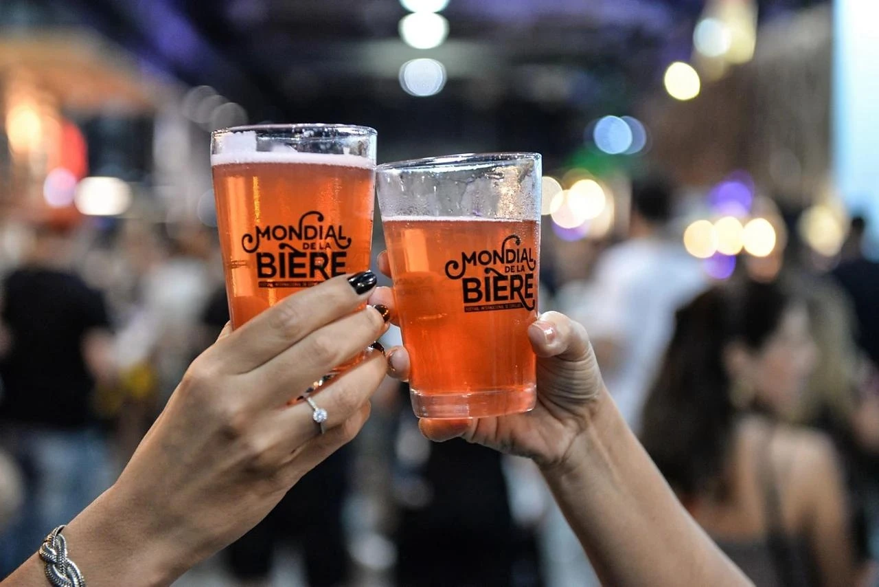 Brinde com dois copos de cerveja do Mondial de la Bière, com o público do festival ao fundo
