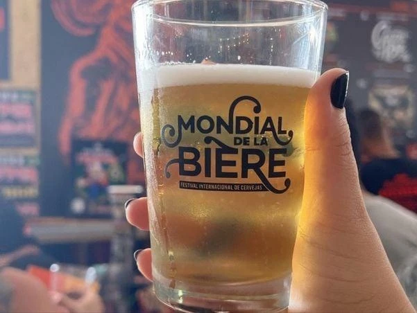 Copo oficial do Mondial de la Bière com cerveja clara, erguido diante de um painel ilustrado
